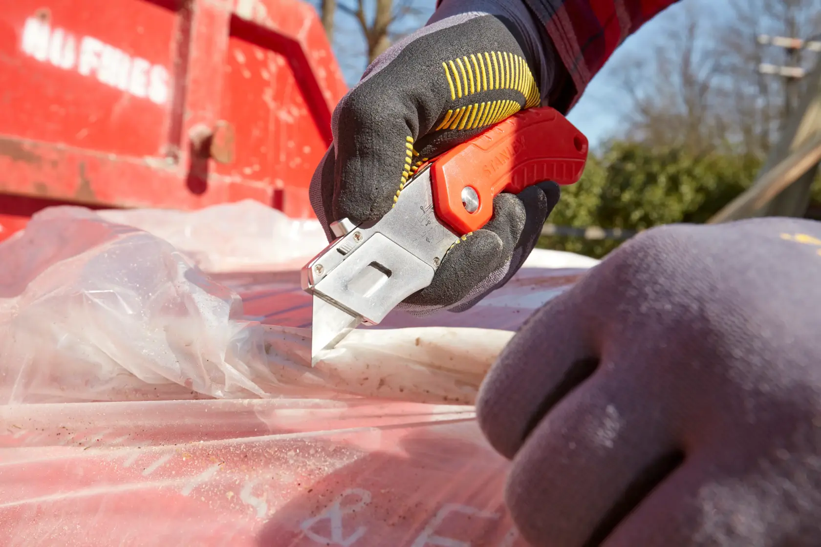 Photo du couteau utilitaire STANLEY 0-10-243 coupant une feuille plastique en extérieur, ouvrier avec gants.