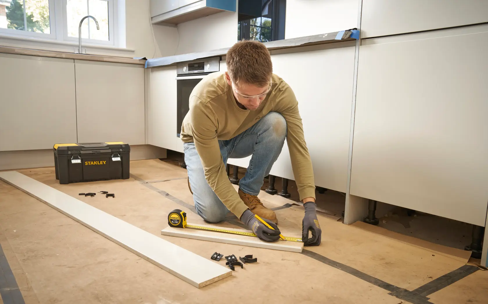 Photo of STANLEY 0-30-656 tape measure used by a person measuring a wooden board in a kitchen, STANLEY toolbox nearby.