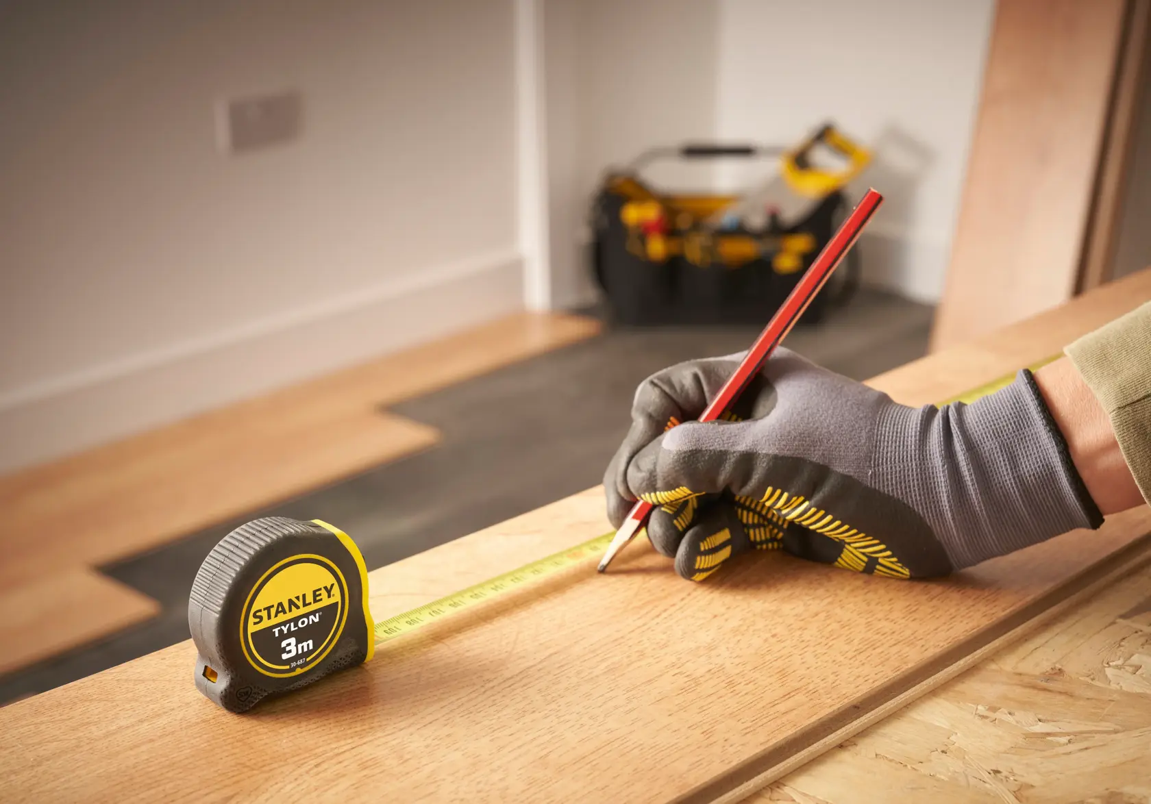 Photo of STANLEY 0-30-687 tape measure on wood, worker with gloves marking with red pencil, tool bag in background.