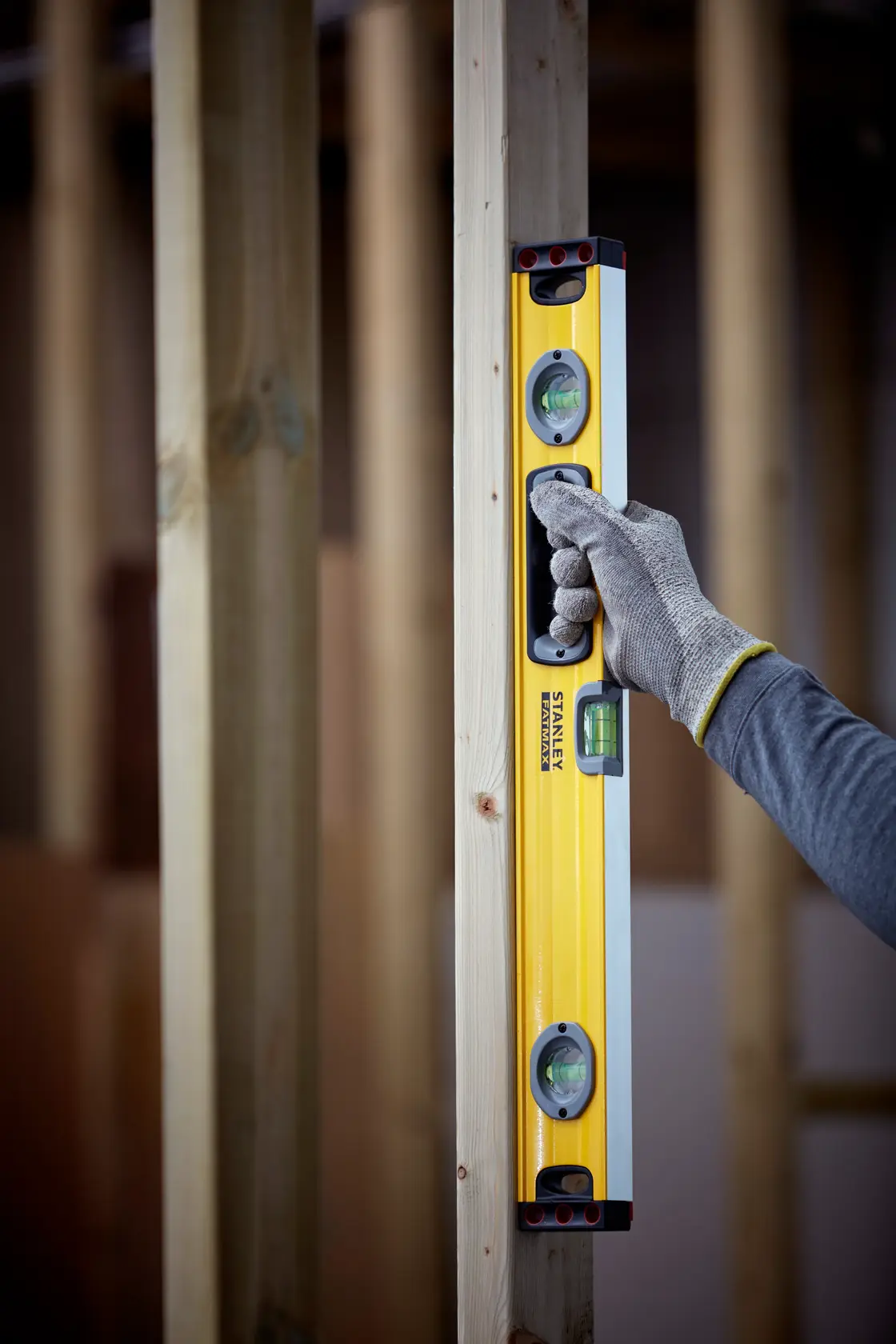 STANLEY 1-43-524 yellow spirit level held vertically against wood post, gloved hand visible, construction setting.