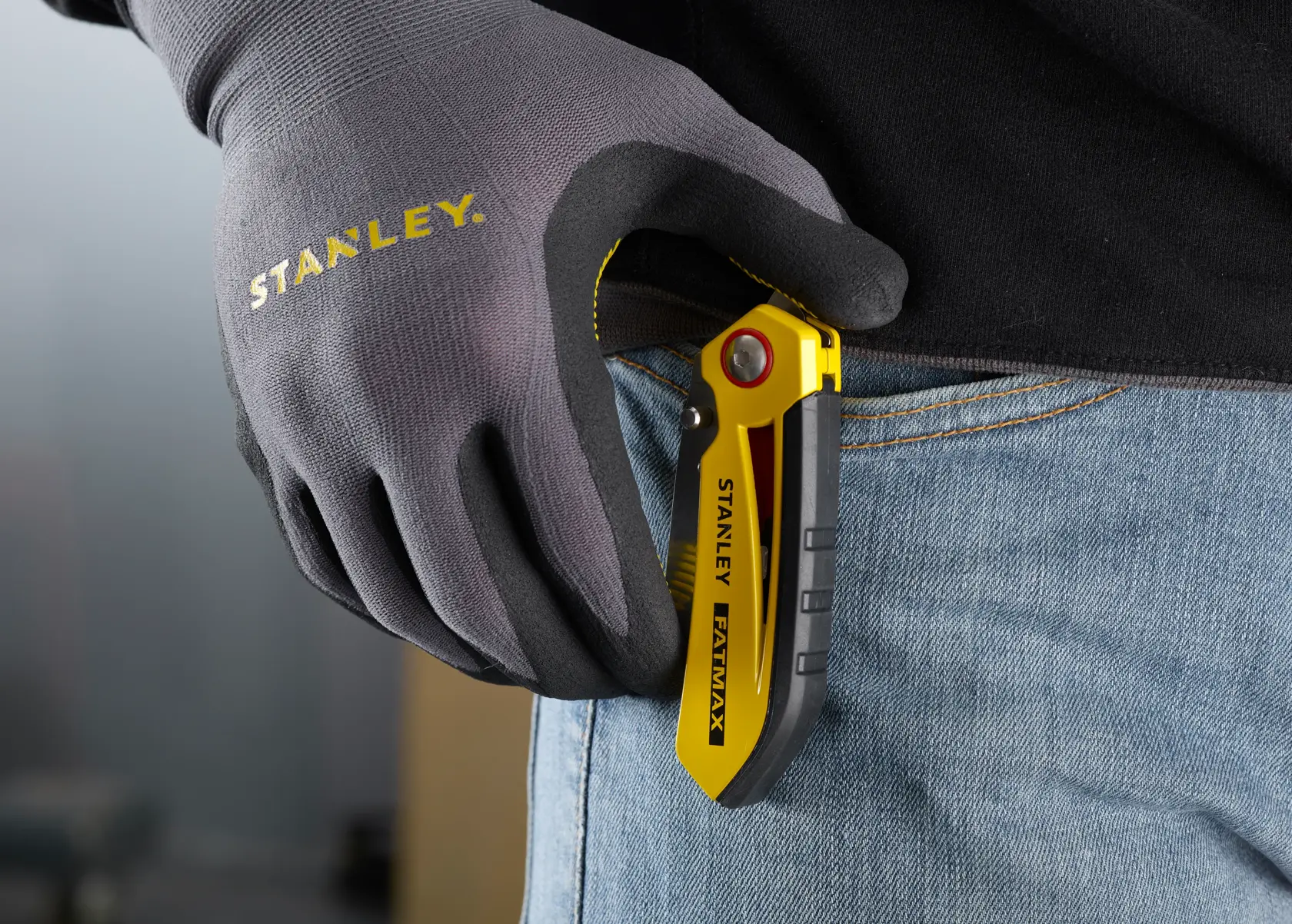 STANLEY FMHT0-10827 folding utility knife in yellow and black clipped to jeans, gloved hand holding it.