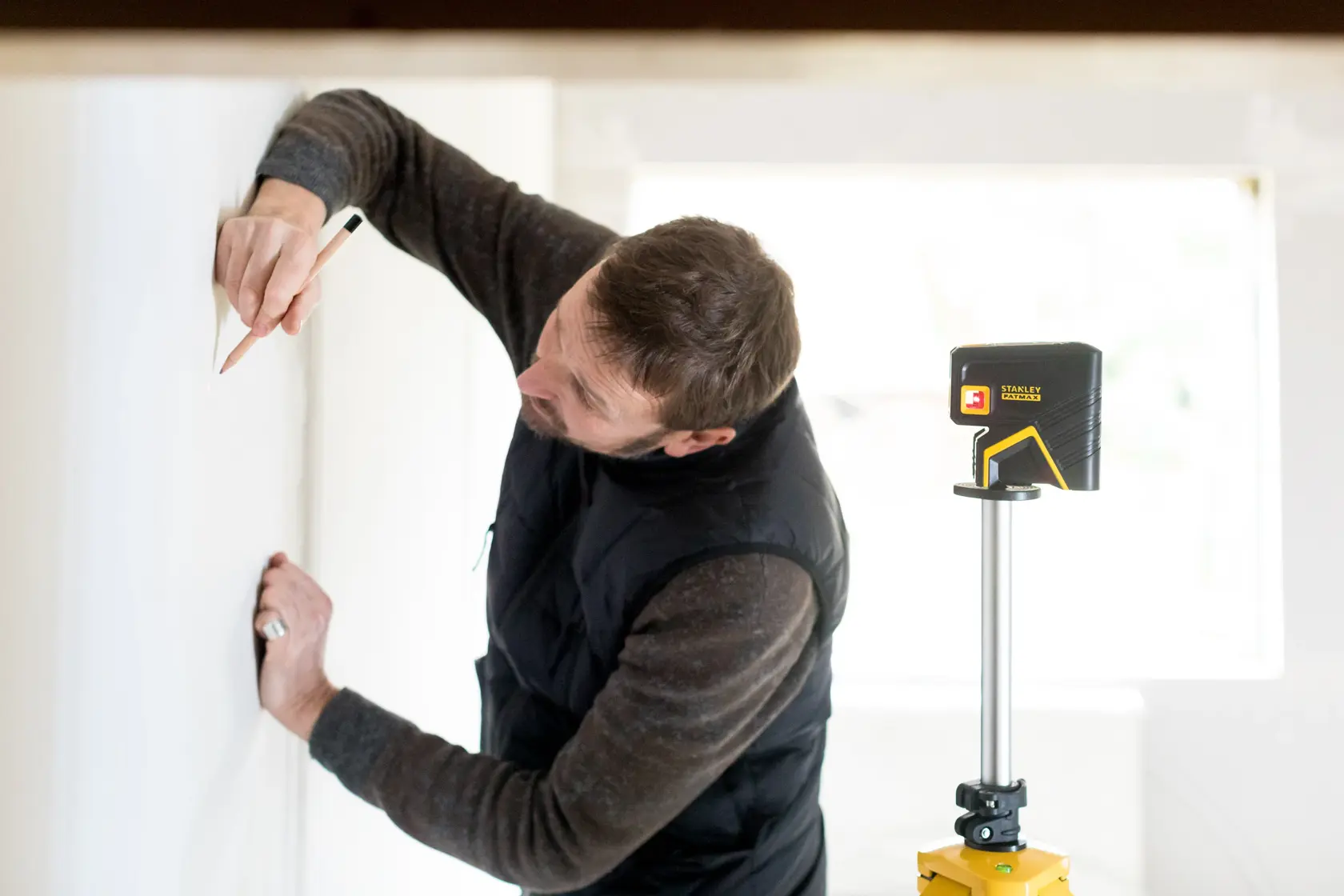 Stanley FMHT1-77413 laser level on tripod beside person marking wall, indoor setting.