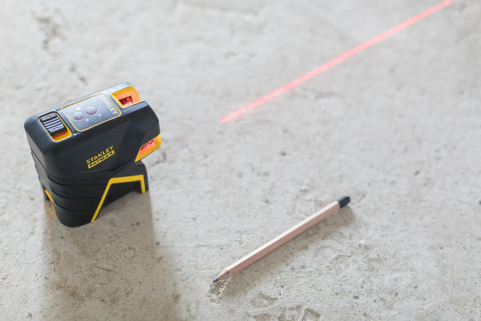 STANLEY FMHT1-77415 cross line laser level with visible red laser line and pencil on concrete floor, angled view.