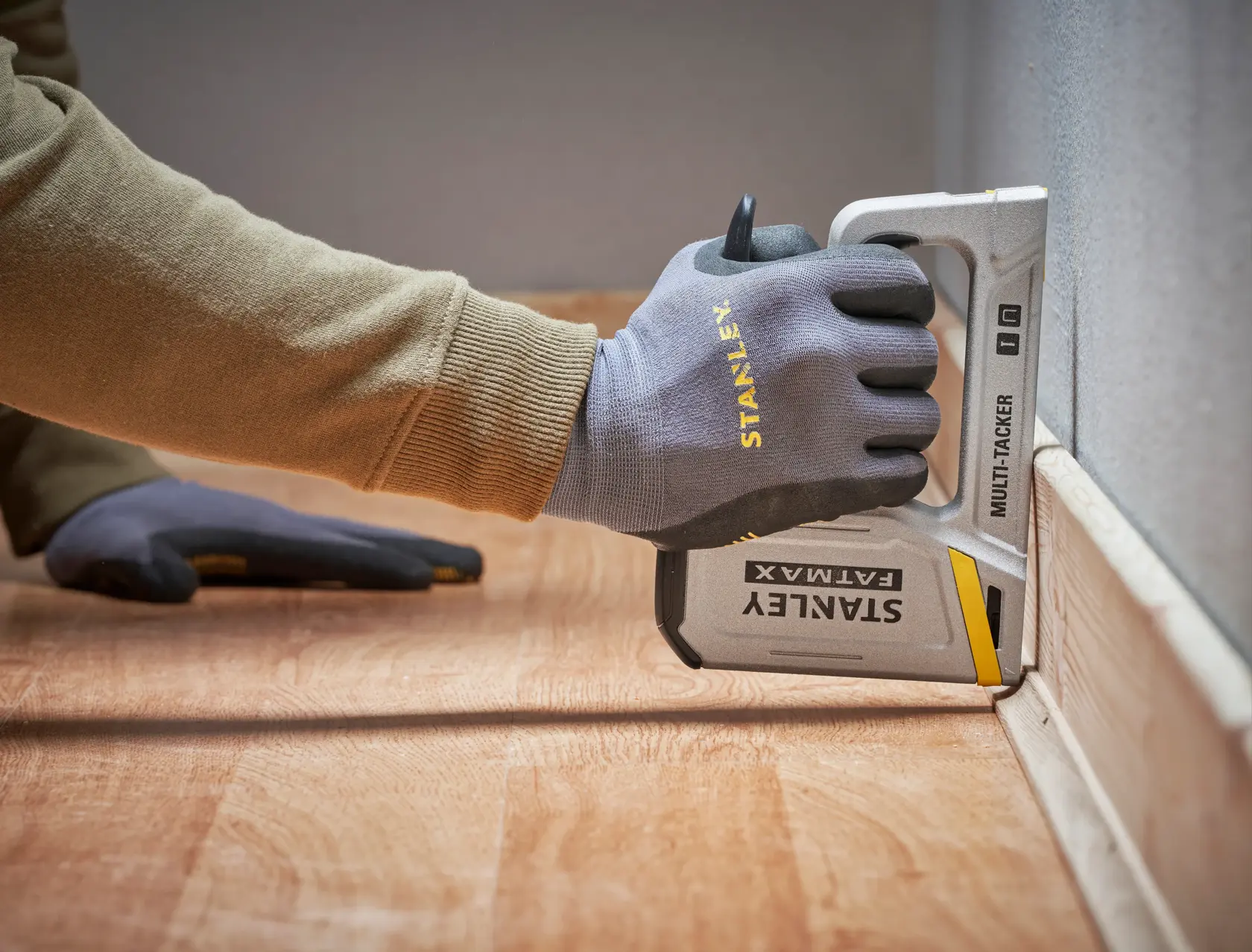 Photo of a gloved hand using a STANLEY FATMAX FMHT70443-0 multi-tacker stapler on flooring next to a wall.