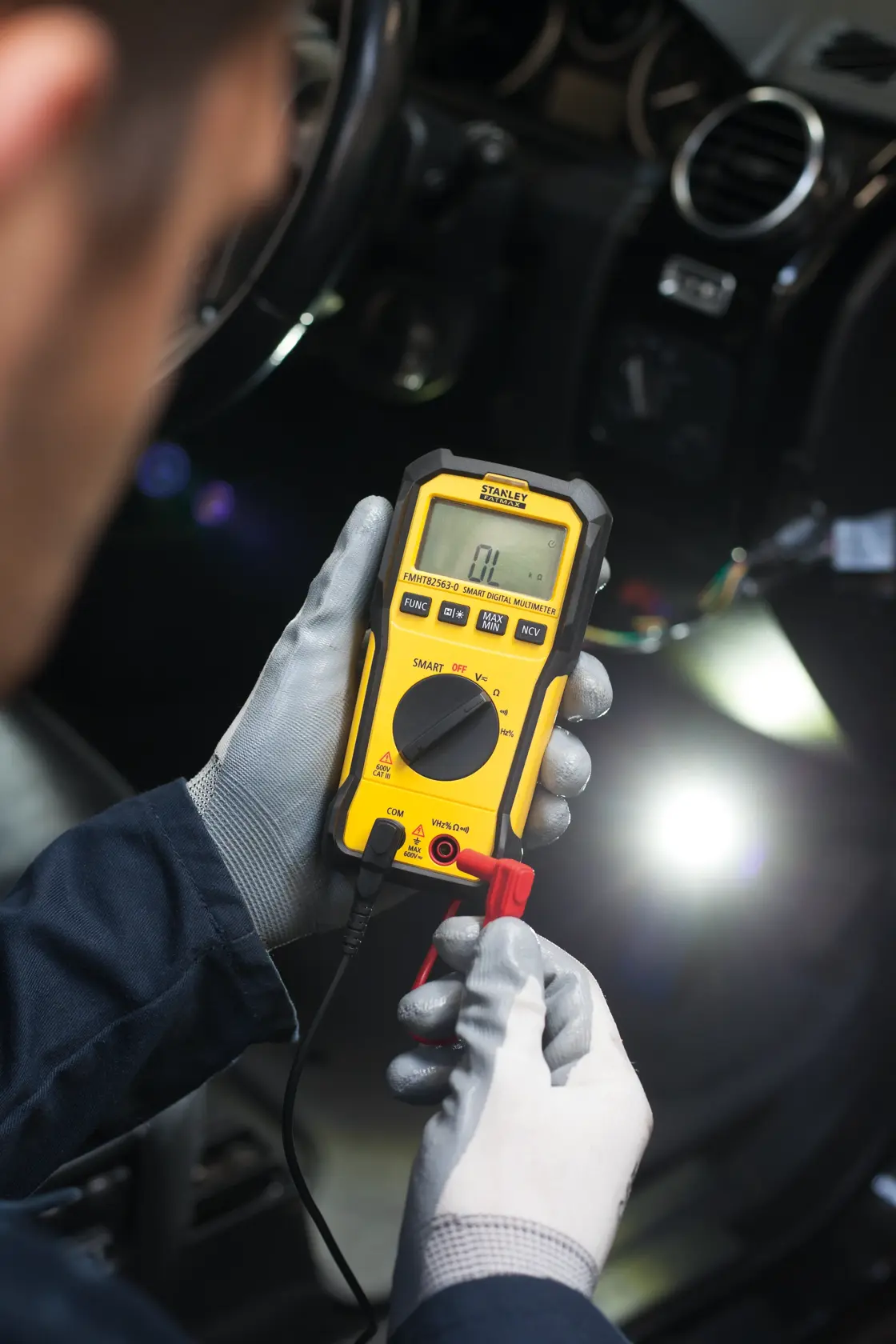 Photo of STANLEY FMHT82563-0 digital multimeter used for car diagnostics, held by gloved hands.