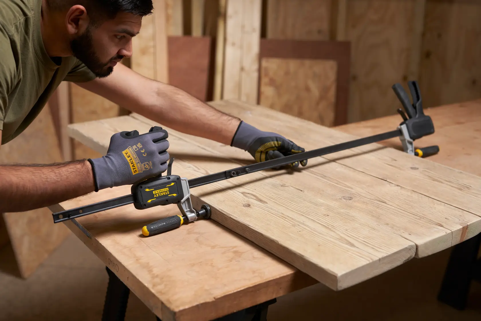 STANLEY FMHT83006-0 bar clamp holding wooden boards on workbench, worker wearing STANLEY gloves.