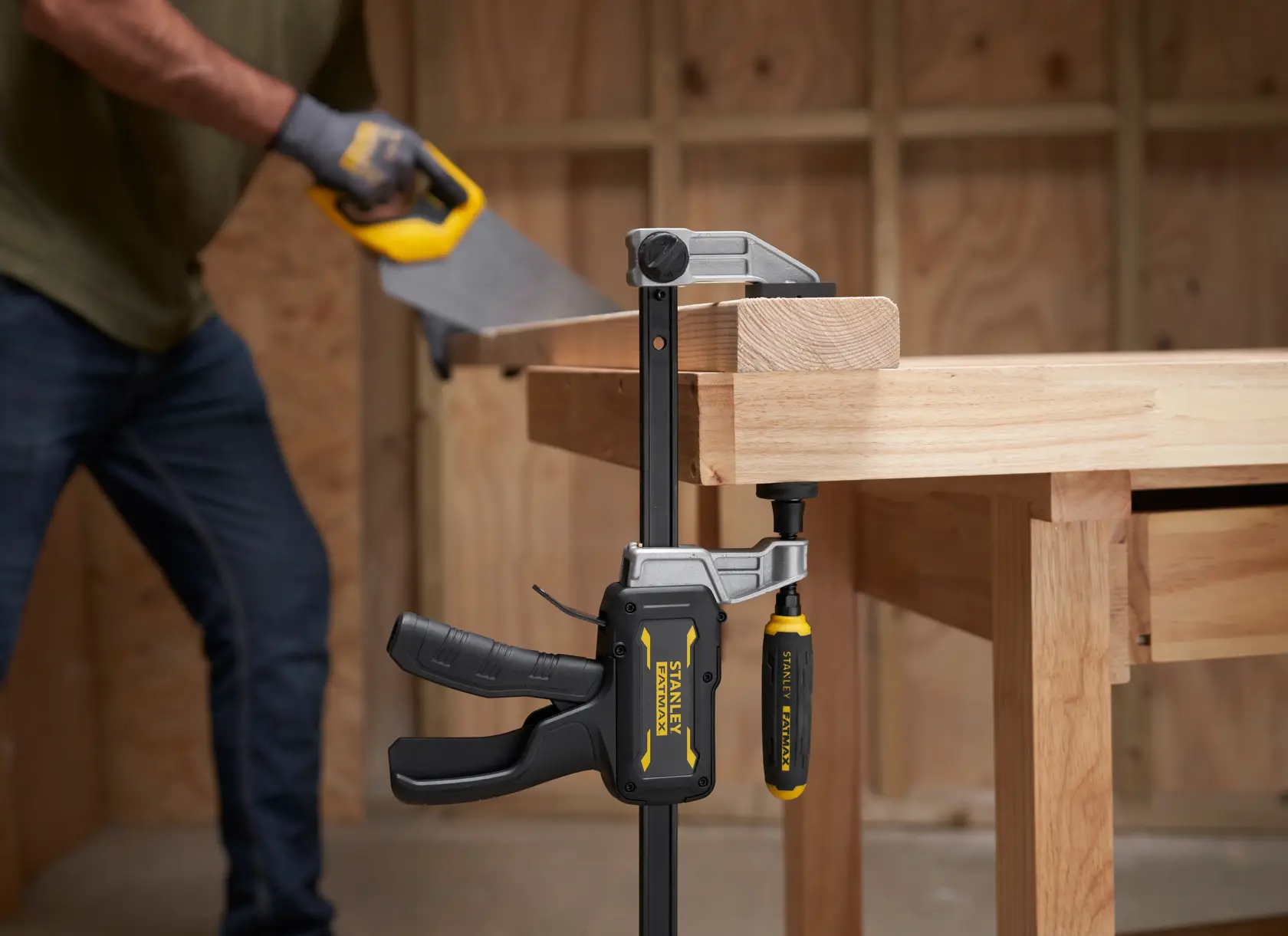 STANLEY FMHT83008-0 clamp securing wood on workbench, person sawing wood in workshop.