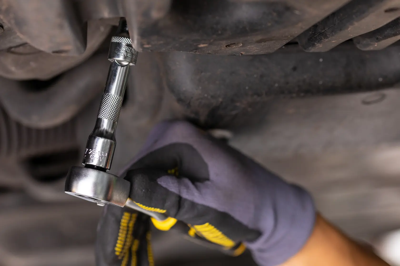 STANLEY FMMT25131-0 ratchet wrench in use under vehicle, close-up photography, tool held by gloved hand.