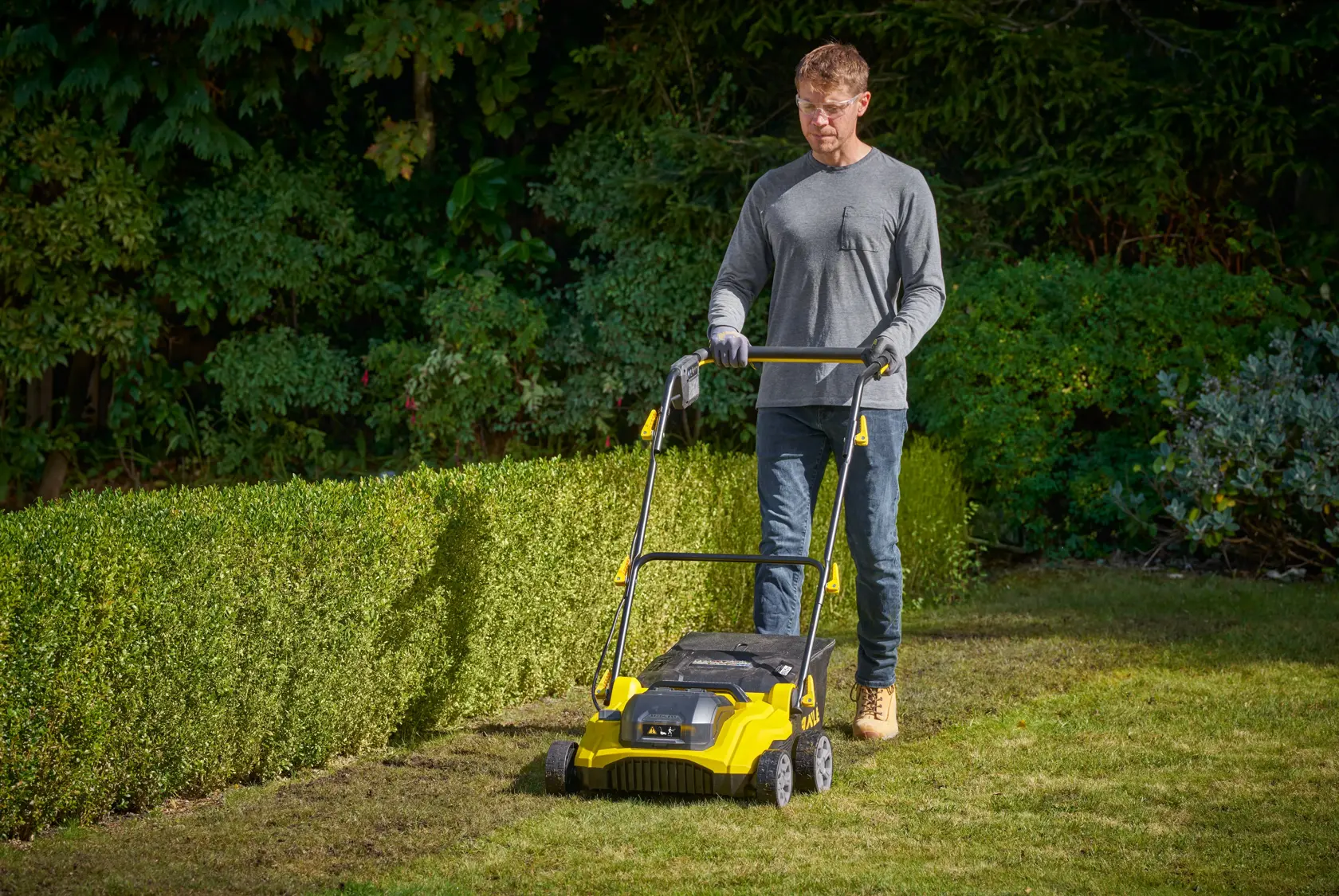 STANLEY SCOEP3801N scarificatore per prato giallo in uso in un giardino, con vegetazione sullo sfondo.