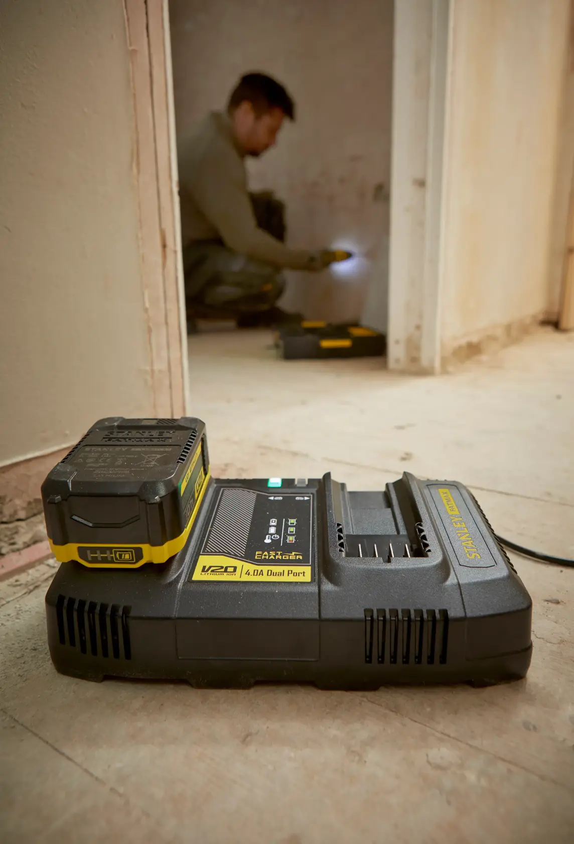 Caricabatterie doppia porta STANLEY SFMCB24 V20 su pavimento dell’officina con batteria collegata, operaio sullo sfondo.