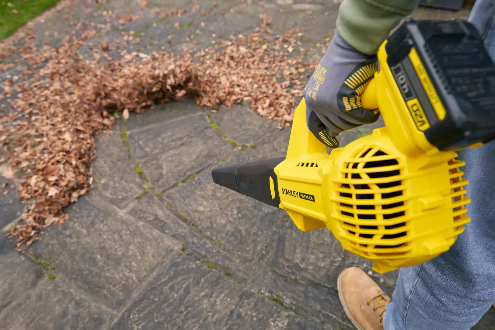 Souffleur de feuilles STANLEY SFMCBL714M1 utilisé sur une terrasse en pierre extérieure, déplaçant un tas de feuilles sèches.