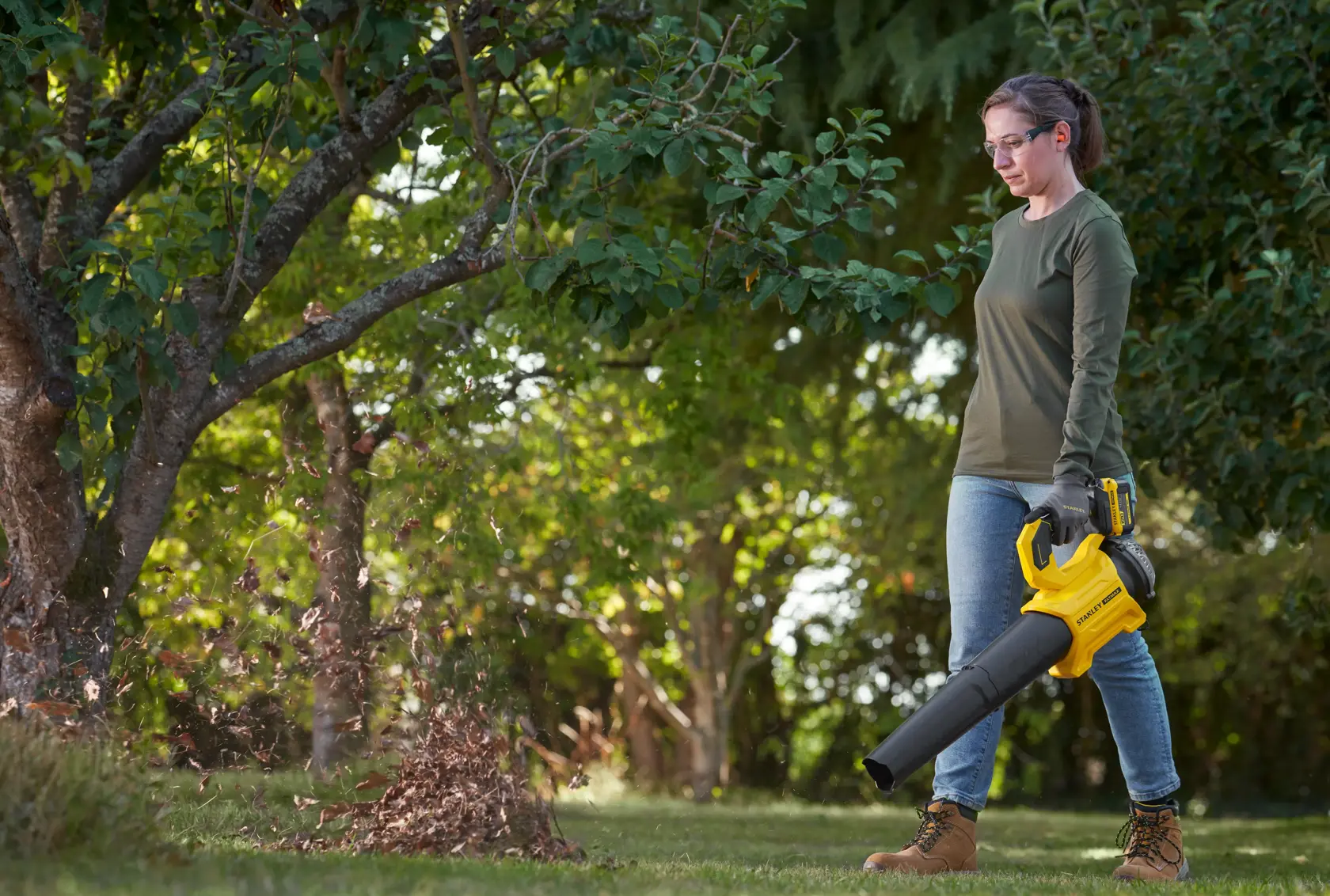 Souffleur de feuilles STANLEY SFMCBL730B utilisé en extérieur, design jaune et noir, déplaçant des feuilles sous un arbre.