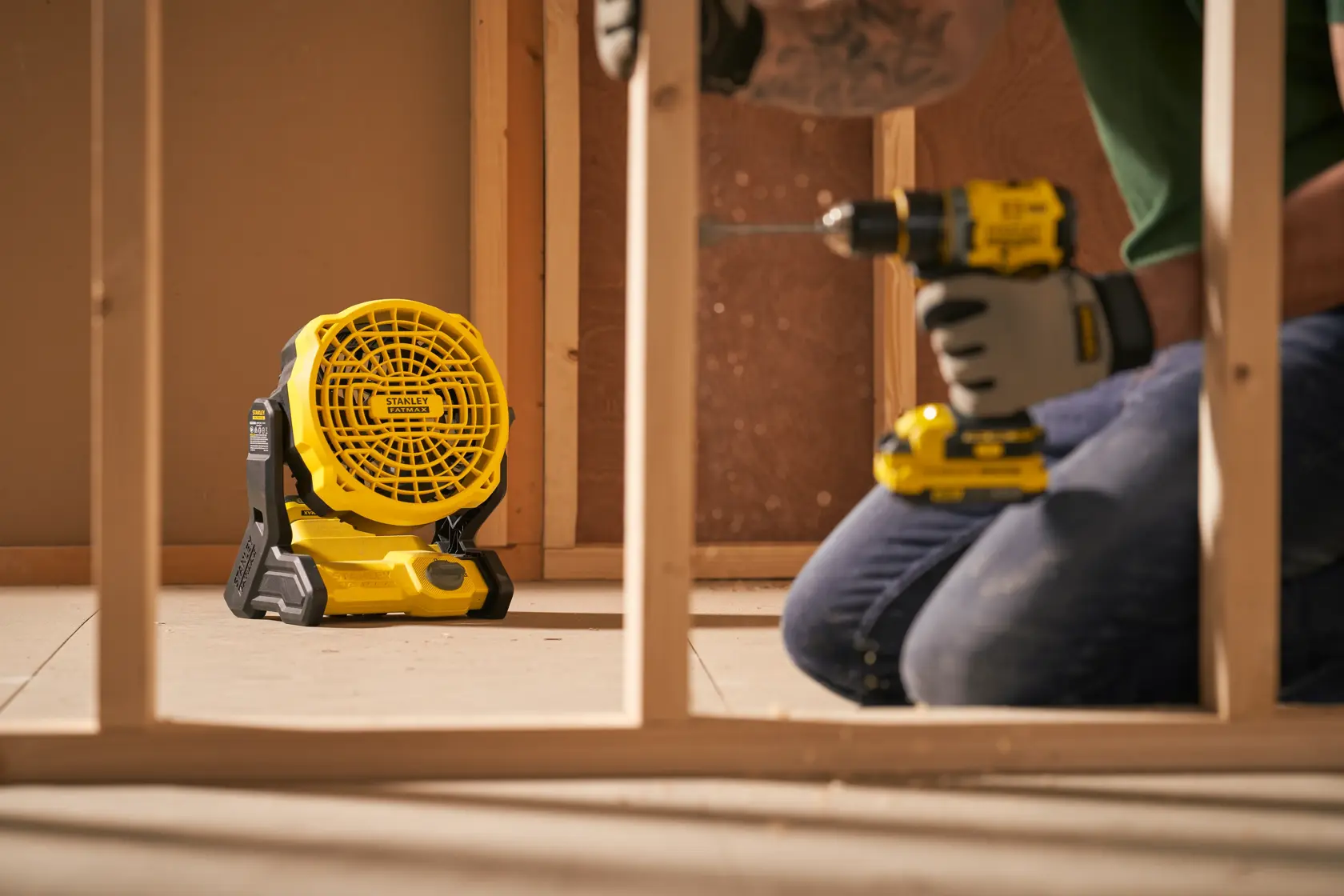 Ventilateur de chantier portable Stanley SFMCE001B au sol dans un chantier, ouvrier utilisant une perceuse à proximité.