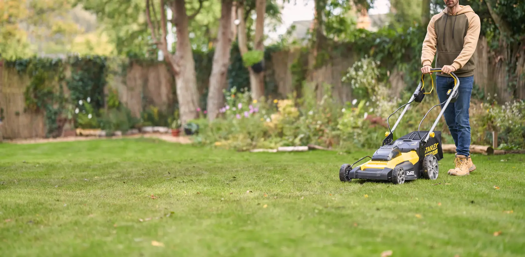Photo of STANLEY SFMCMWS251M lawn mower in use on grass, yellow and black design, garden background.