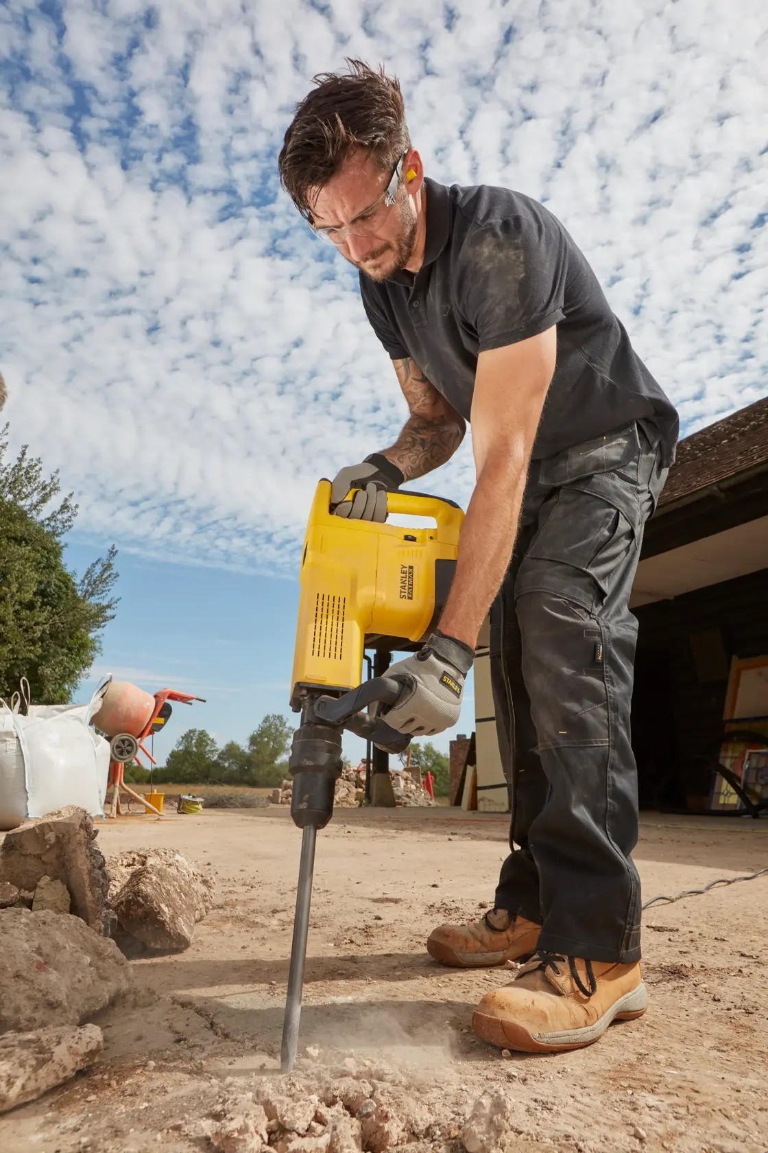 Photo of a person using STANLEY SFMEH230K-QS demolition hammer on outdoor concrete, construction scene visible.