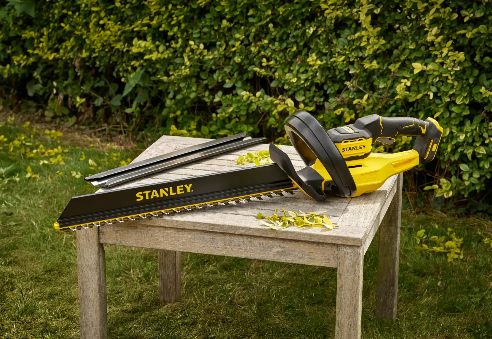 STANLEY STCHT8255B taille-haie jaune et noir sur une table en bois à l'extérieur, lames et feuilles visibles.