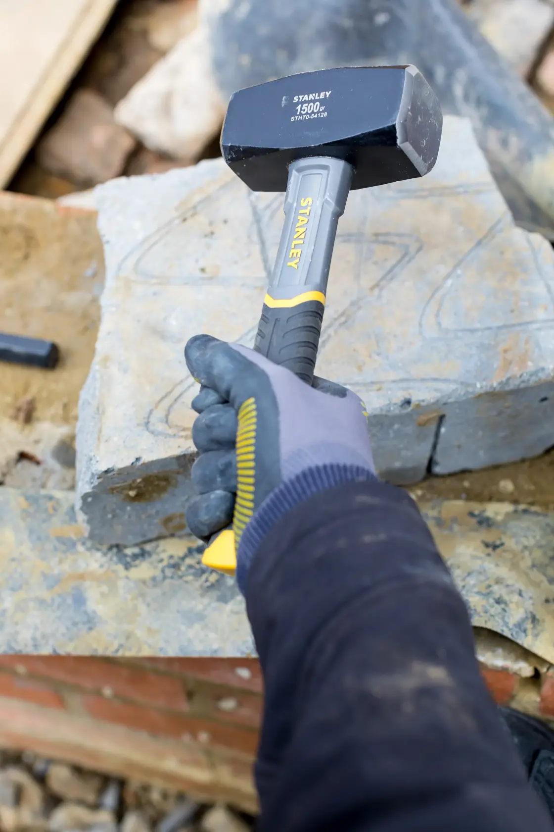 STANLEY STHT0-54128 sledge hammer with yellow and black handle held by gloved hand on stone construction site.