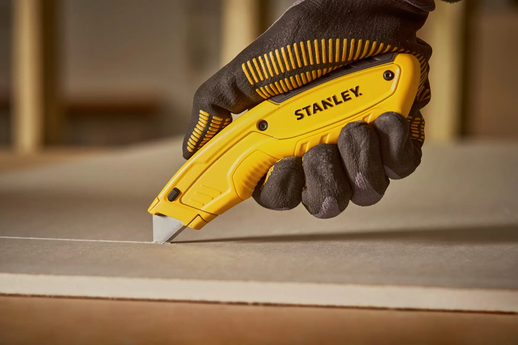 Photo of STANLEY STHT10430-0 utility knife cutting drywall, held by gloved hand.
