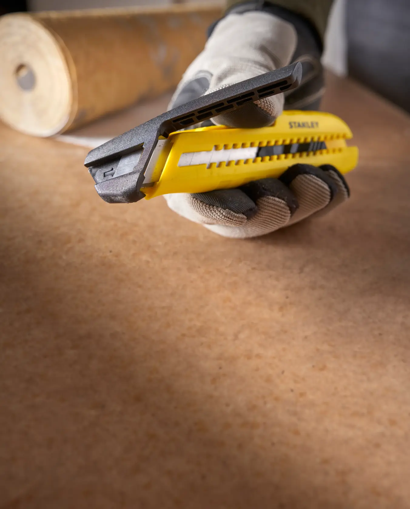 Foto des gelben STANLEY STHT10489 Universalmessers in behandschuhter Hand auf Arbeitsfläche, Materialrolle im Hintergrund.