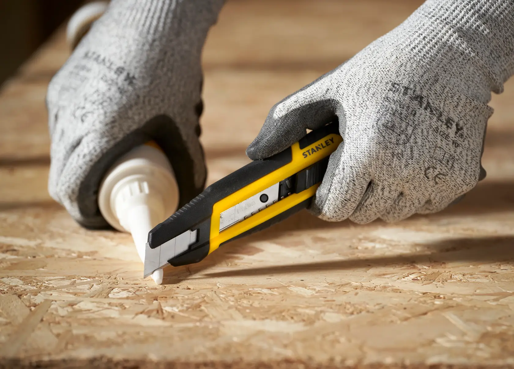 Photo of STANLEY STHT10501-0 utility knife cutting on a wooden surface, held by gloved hands.