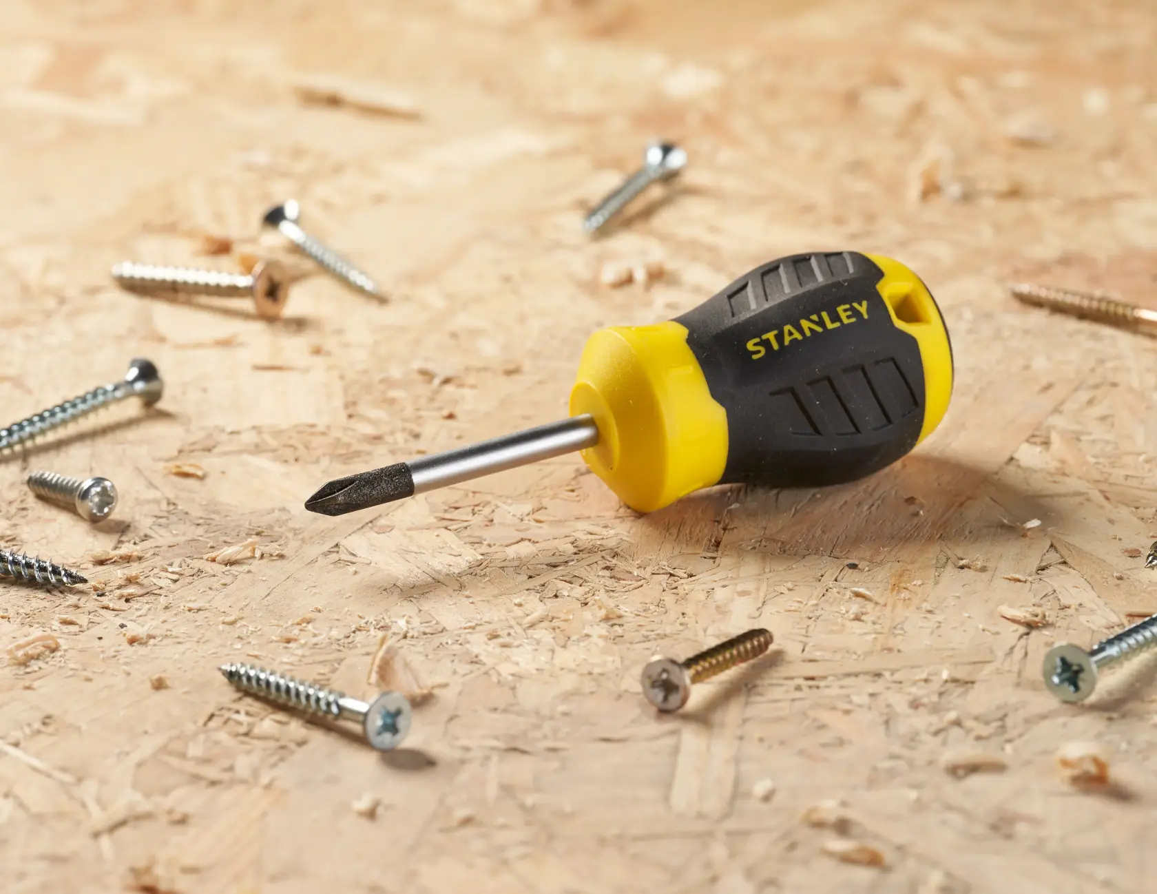 STANLEY STHT16154-0 stubby Phillips screwdriver with black and yellow handle on wood surface with scattered screws.