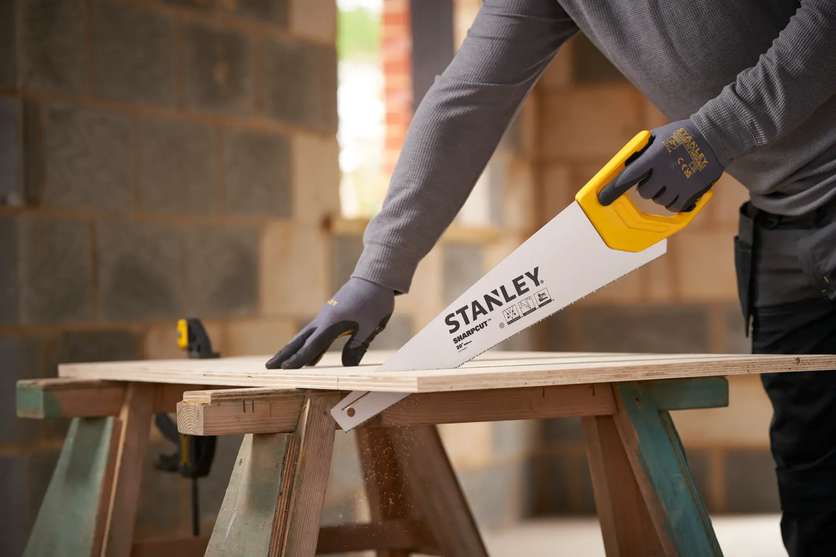Scie à main Stanley STHT20375-1 avec poignée jaune découpant une planche de bois sur tréteaux, ouvrier ganté.