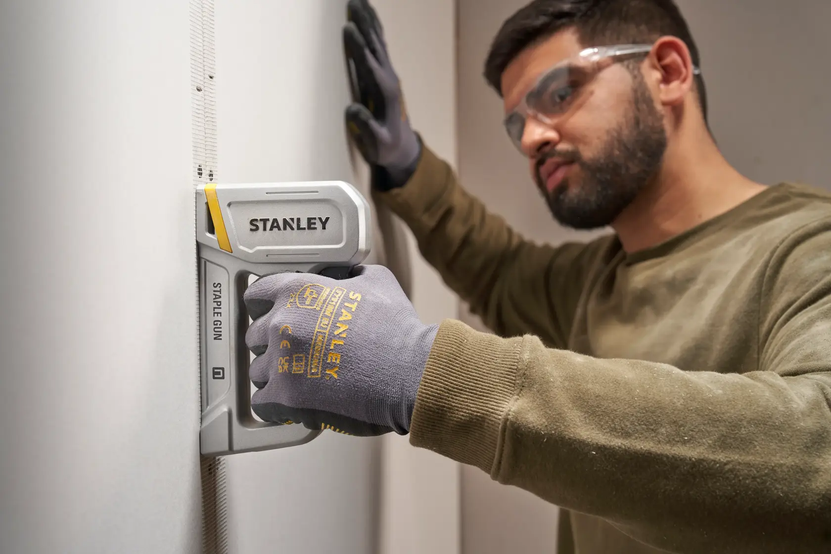 Photo of STANLEY STHT70150-0 staple gun used on wall, person wearing STANLEY gloves holding the tool.