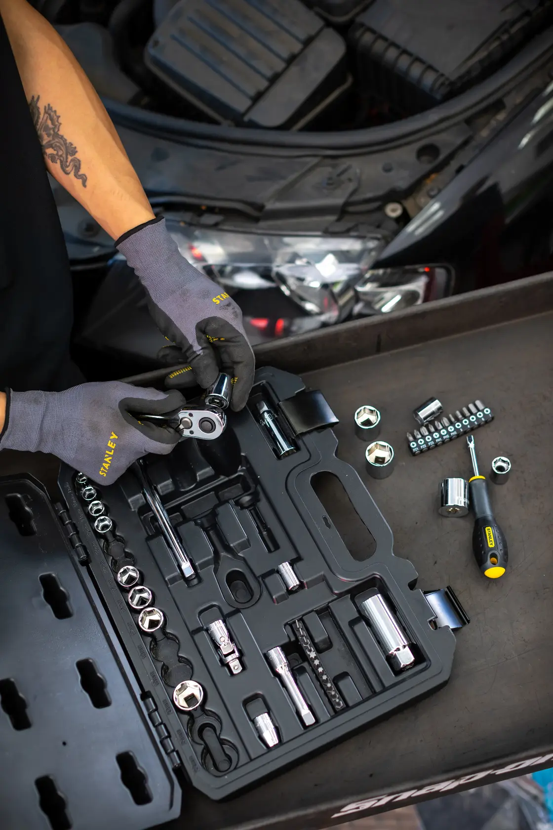 STANLEY STMT82829-1 mechanics tool set open on a table, gloves, car engine in background.